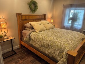 A bedroom with a wooden bed frame, a green patterned bedspread, a wooden nightstand with a lamp, and a window with a view.