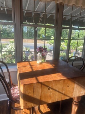 Una mesa de madera con un jarrón de flores, dos sillas y vistas a un jardín a través de amplias ventanas.