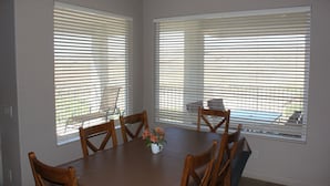 Un comedor con una mesa y sillas, ventanas amplias con persianas y un pequeño jarrón con flores sobre la mesa.