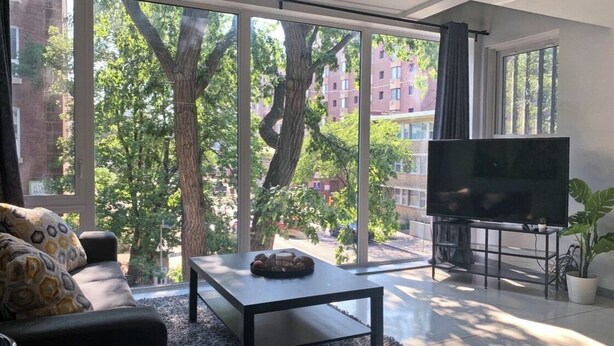 Un salon moderne doté d’un grand téléviseur, d’un canapé, d’une table basse et offrant une vue sur les arbres et les bâtiments grâce à de grandes fenêtres.