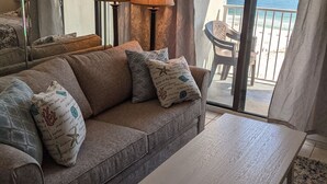 A living room with a sofa, pillows, a table, and a view of the beach through large windows. - Beautiful Gulf Front Unit with Balcony (Gulf Shores)