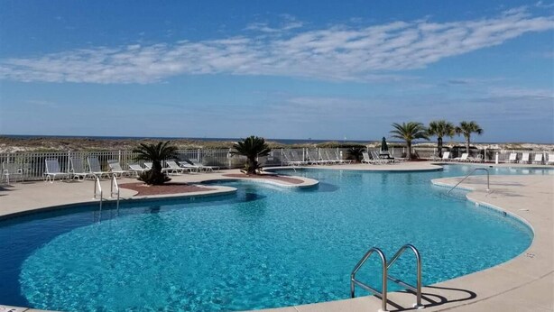 A large, clear swimming pool with a curved design, surrounded by lounge chairs and palm trees, with a view of the beach and ocean in the background. - Beautiful Gulf Front Unit with Balcony (Gulf Shores)