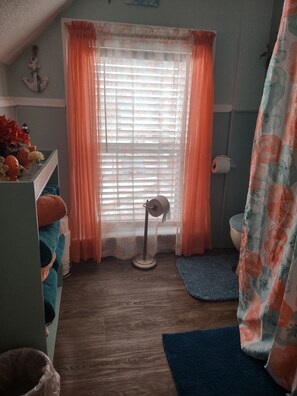 Een kamer met een houten vloer, een raam met oranje gordijnen, een staande ventilator, een blauw vloerkleed en een klein plankje met decoratieve objecten.