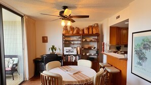 A dining area with a round table, chairs, and a ceiling fan. There is a bookshelf with various items and a kitchen area in the background.
