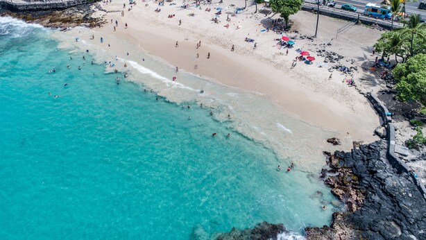 An aerial view of a beach with clear turquoise waters, a sandy shore, and a stone wall.