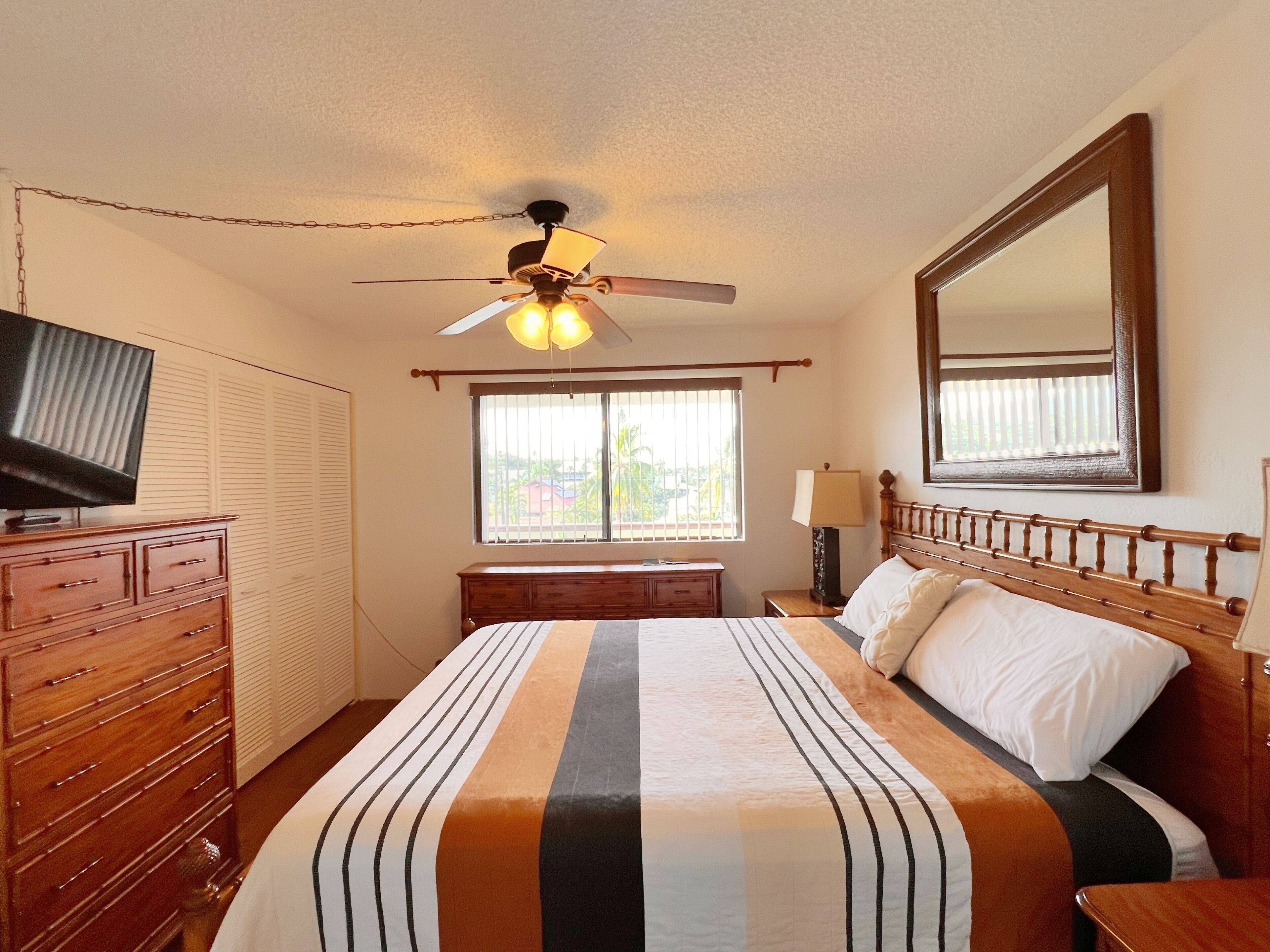 Quarto com cama, cómoda de madeira, televisão, ventoinha no teto, janela e espelho na parede.