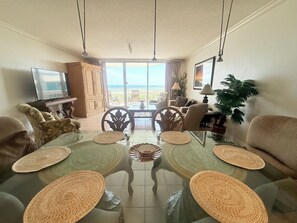 Ein Wohnzimmer mit Esstisch aus Glas, Sofas, einem Fernseher und Blick aufs Meer.