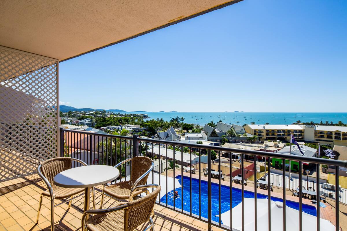 A balcony with wicker chairs and a table overlooking a swimming pool and a coastal view.