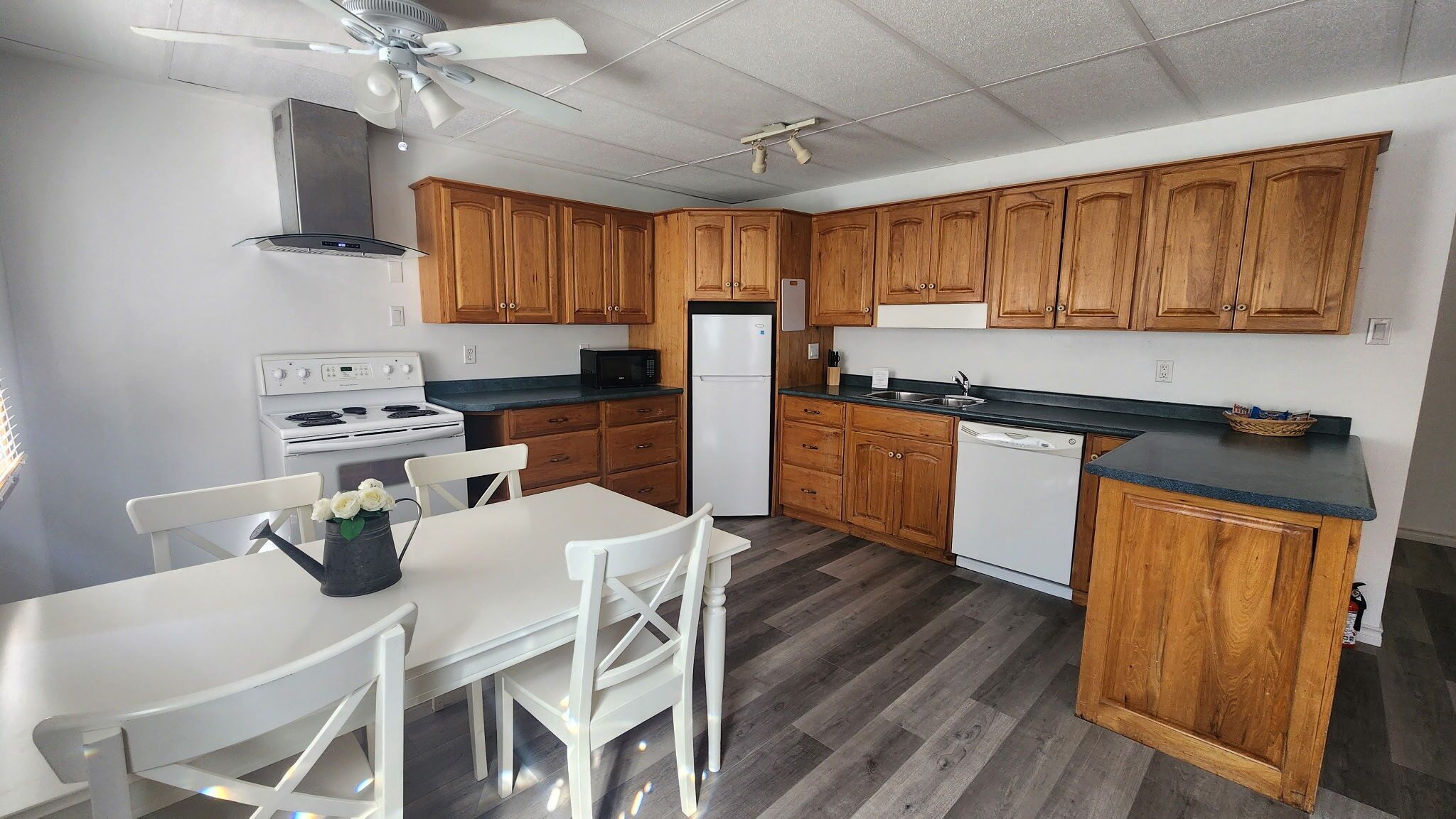 Une cuisine compacte avec des meubles en bois, une cuisinière blanche, un lave-vaisselle et un coin repas comprenant des chaises et une table blanches.
