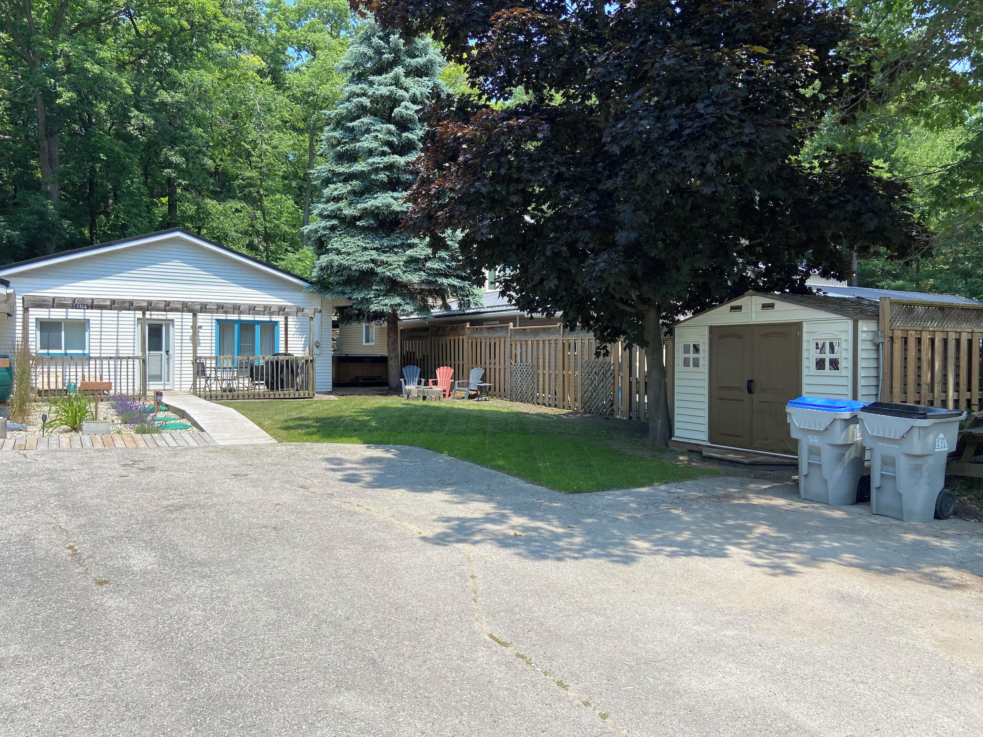 A residential area with a single-story house, a garage, and a small shed. There are two trash bins and a lawn with a few chairs.