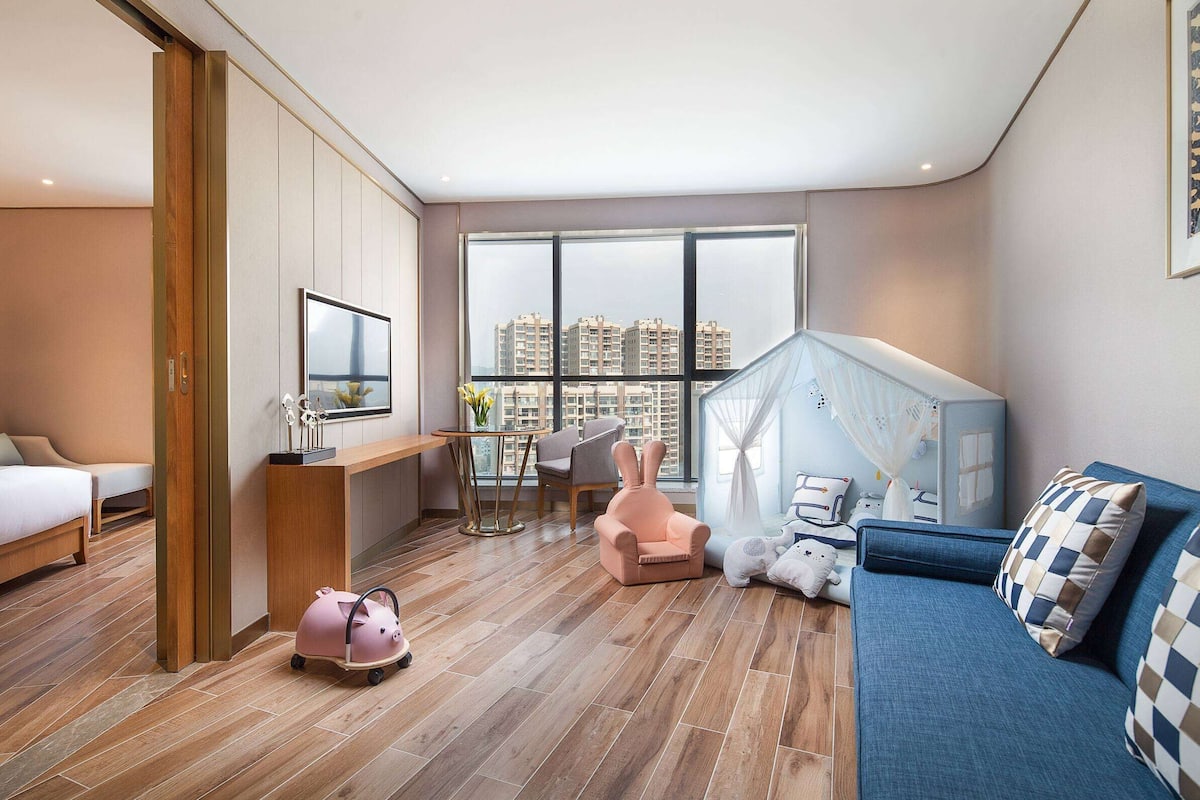 Een moderne hotelkamer met een groot raam, een bed, een bureau, een blauwe bank en een roze fauteuil.