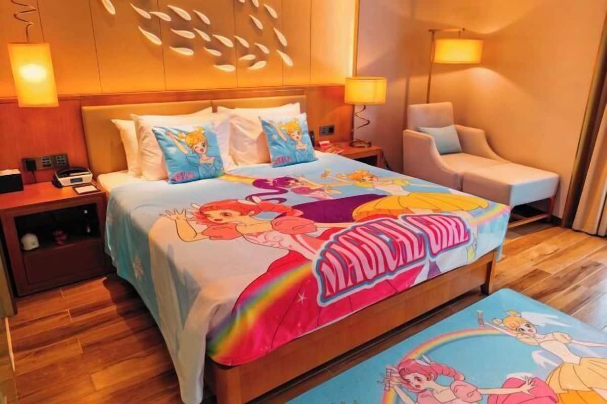 Een hotelkamer met een bed met een sprei met Disney-thema, een nachtkastje met een lamp, een stoel en een houten vloer.