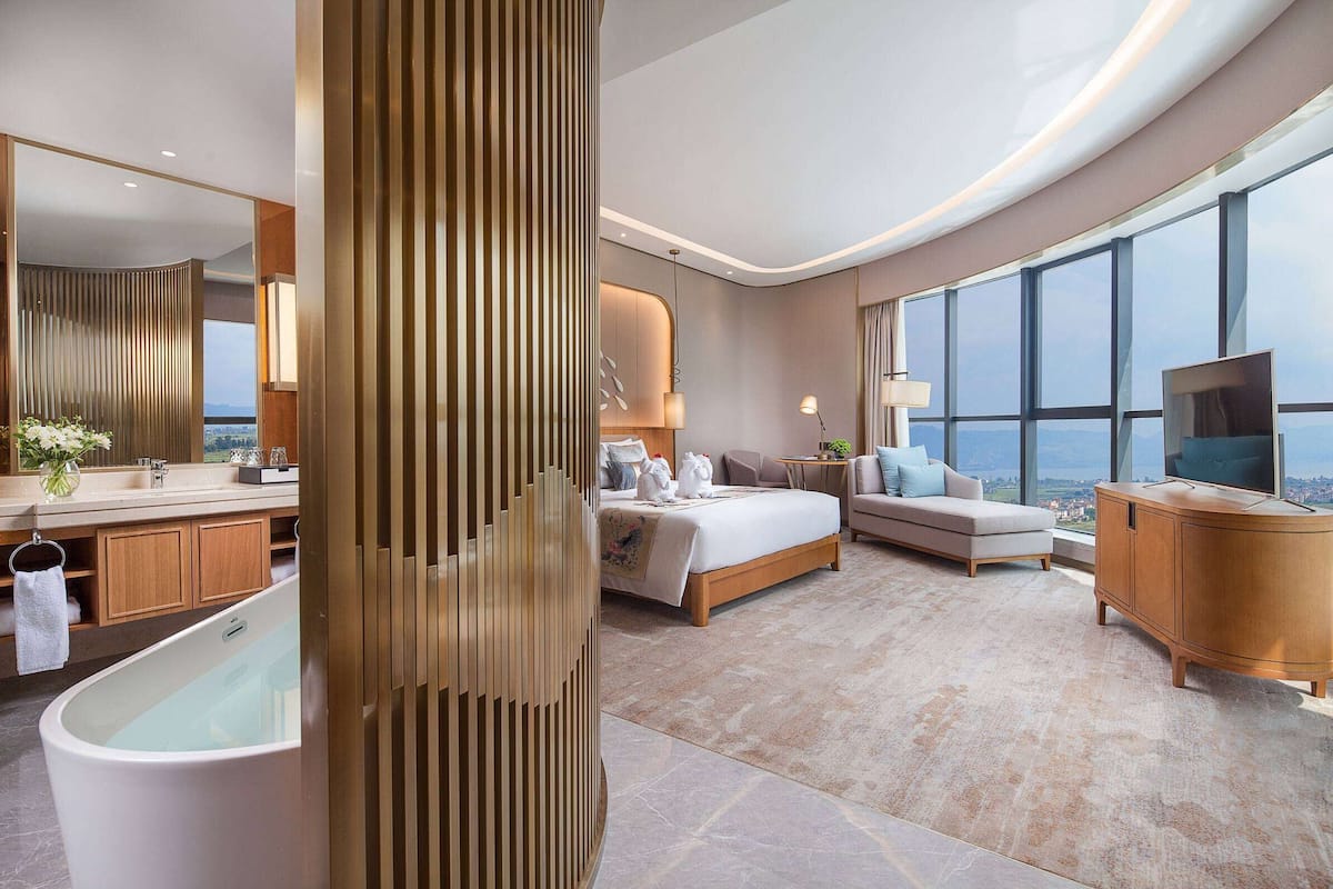 Een moderne hotelkamer met een groot bed, een zithoek, een wastafel met een badkuip en een groot raam met uitzicht op de stad.