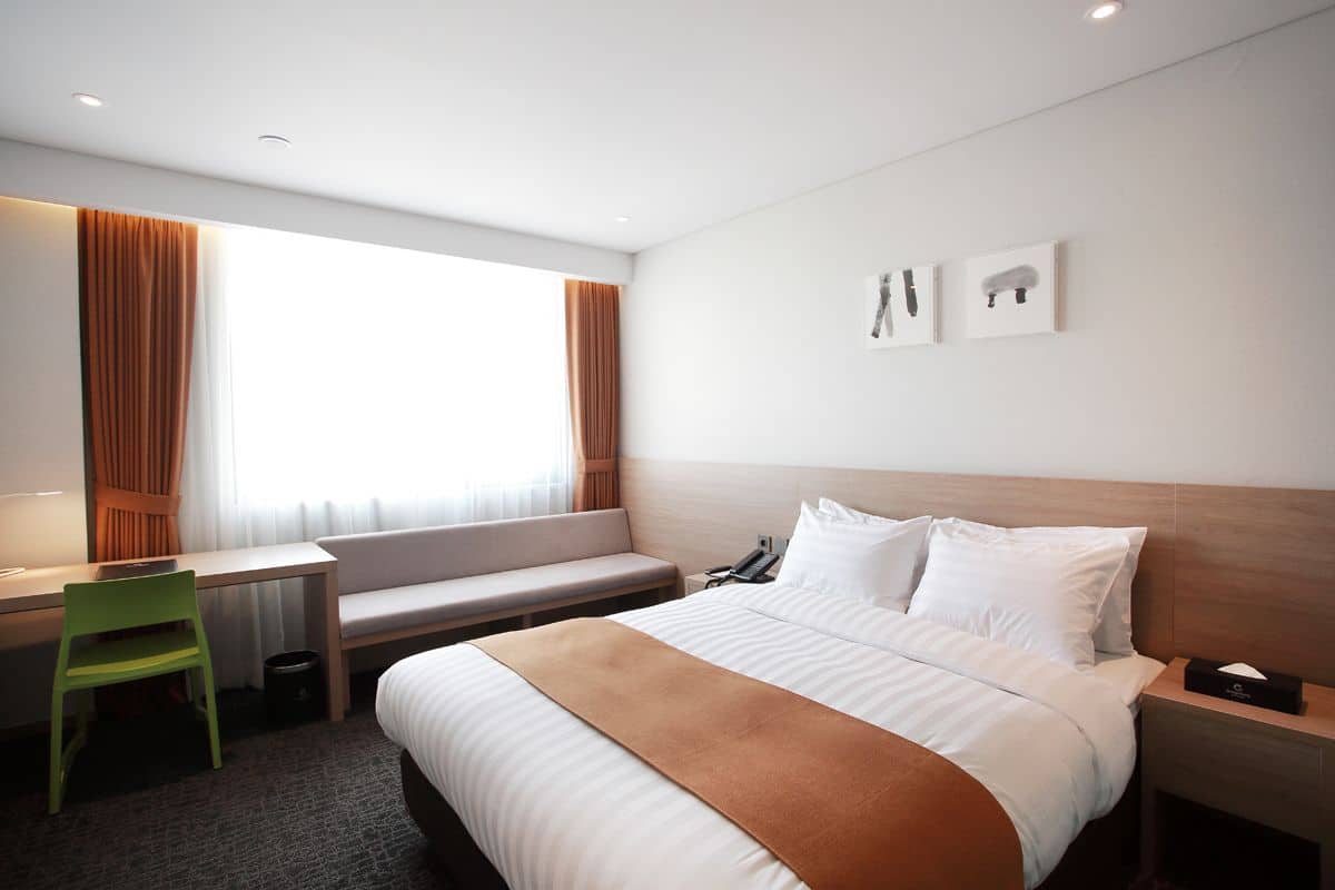 Una habitación de hotel con una cama, un escritorio, una silla, un banco y una ventana con cortinas.