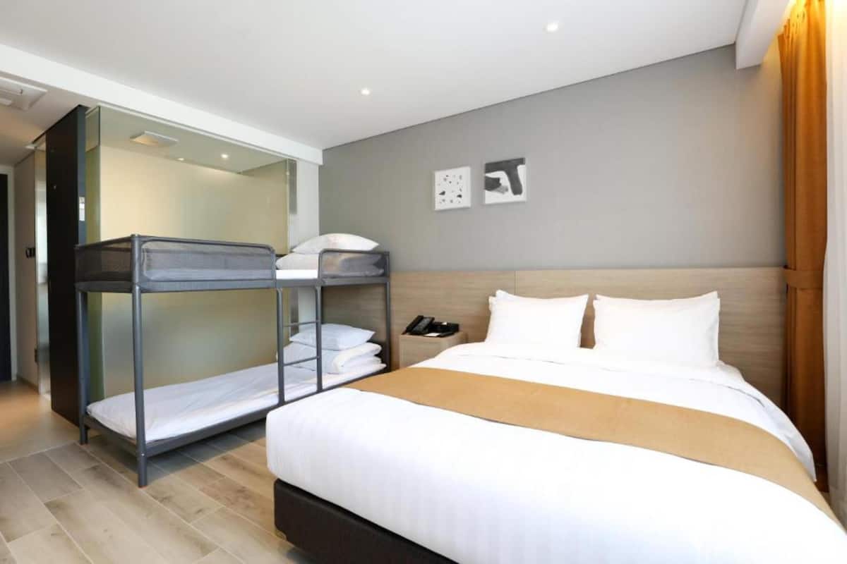 Una habitación de hotel con una cama litera, una cama individual, una mesita de noche con teléfono y una obra de arte colgada en la pared.