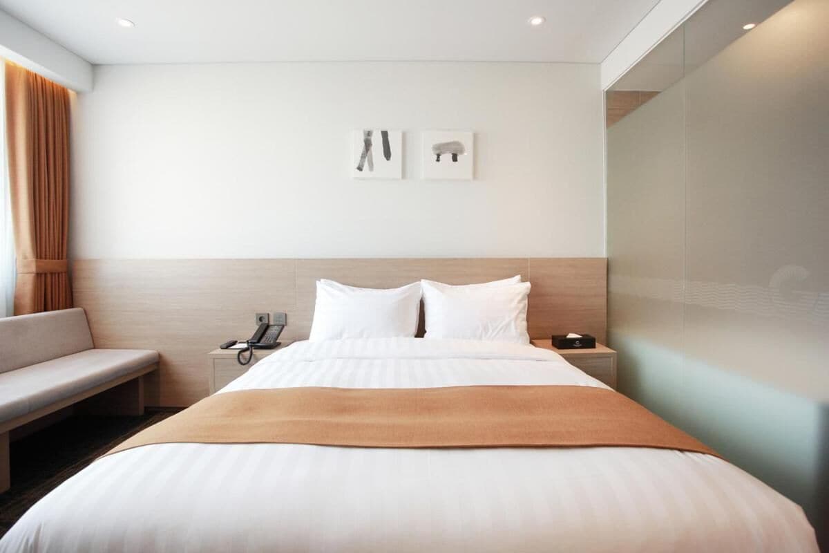 A hotel room with a large bed, a bedside table with a phone, a bench, and two framed pictures on the wall.