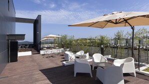An outdoor terrace with white chairs and tables, large umbrellas, and a view of a river and buildings in the background. - Gangneung City Hotel (Gangneung)
