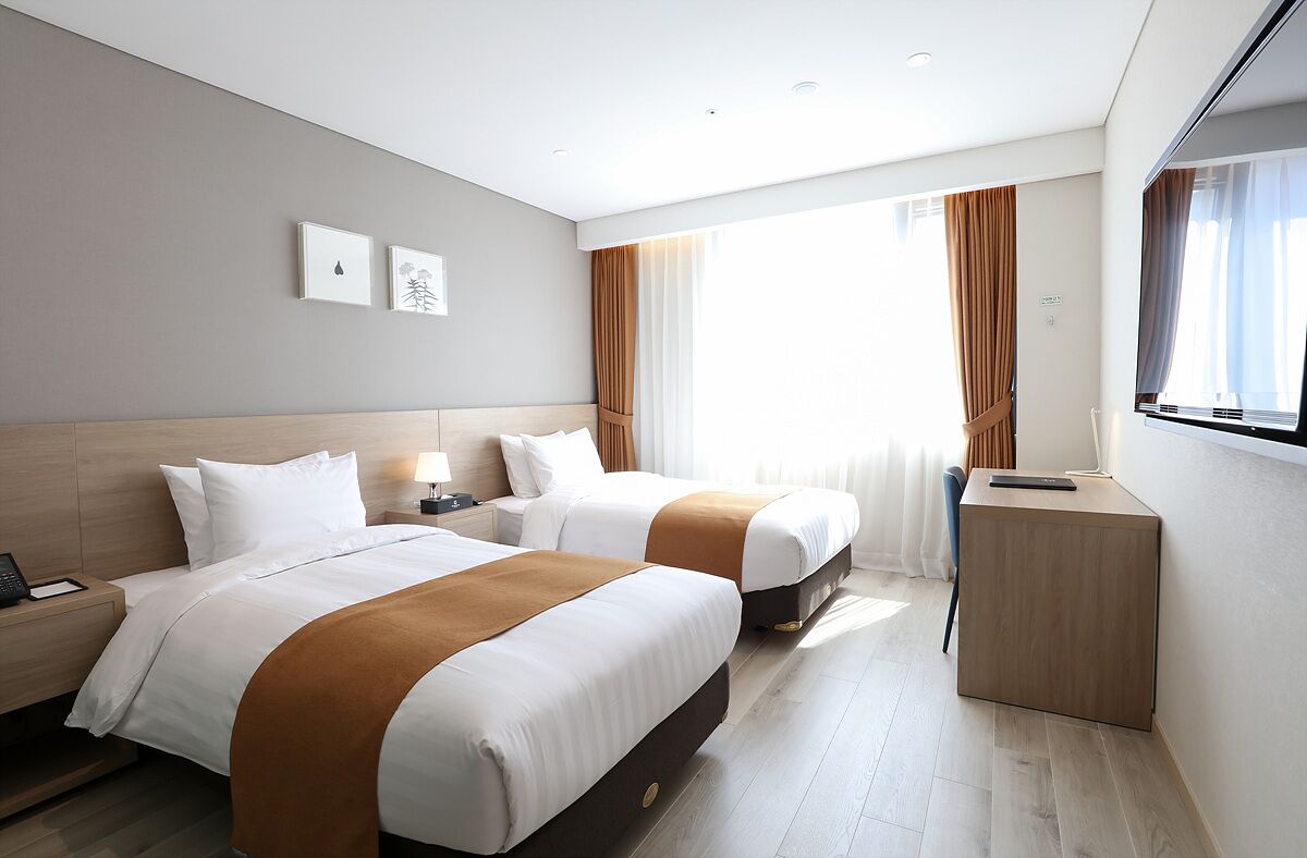 A hotel room with two beds, a bedside table with a lamp, a TV, and a window with curtains.