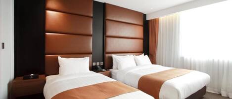 Habitación de hotel con dos camas, un cabecero moderno, una mesita de noche con lámpara y un ventanal con cortinas.