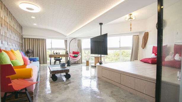 A spacious living area with a large window, a sofa with colorful cushions, a hanging chair, and a flat-screen TV.