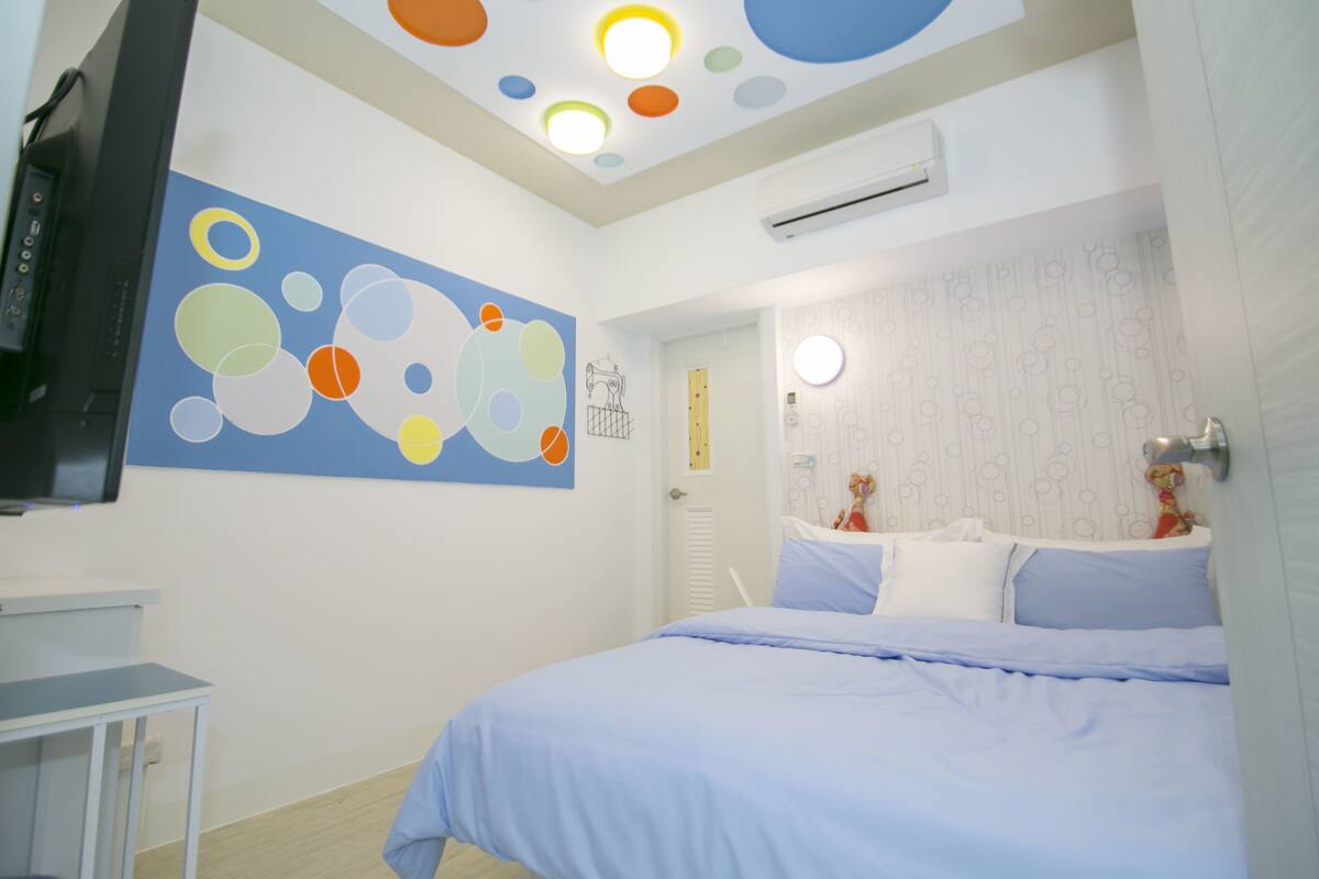 A bedroom with a bed, a wall-mounted TV, a ceiling with circular lights, and a wall with a colorful abstract painting.