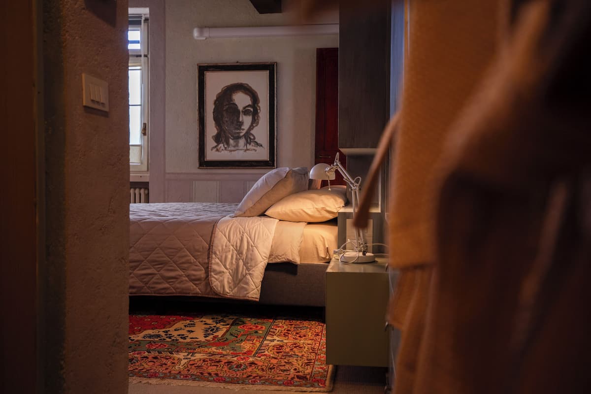 A bedroom with a bed, a bedside table, a lamp, a framed picture on the wall, and a patterned rug.