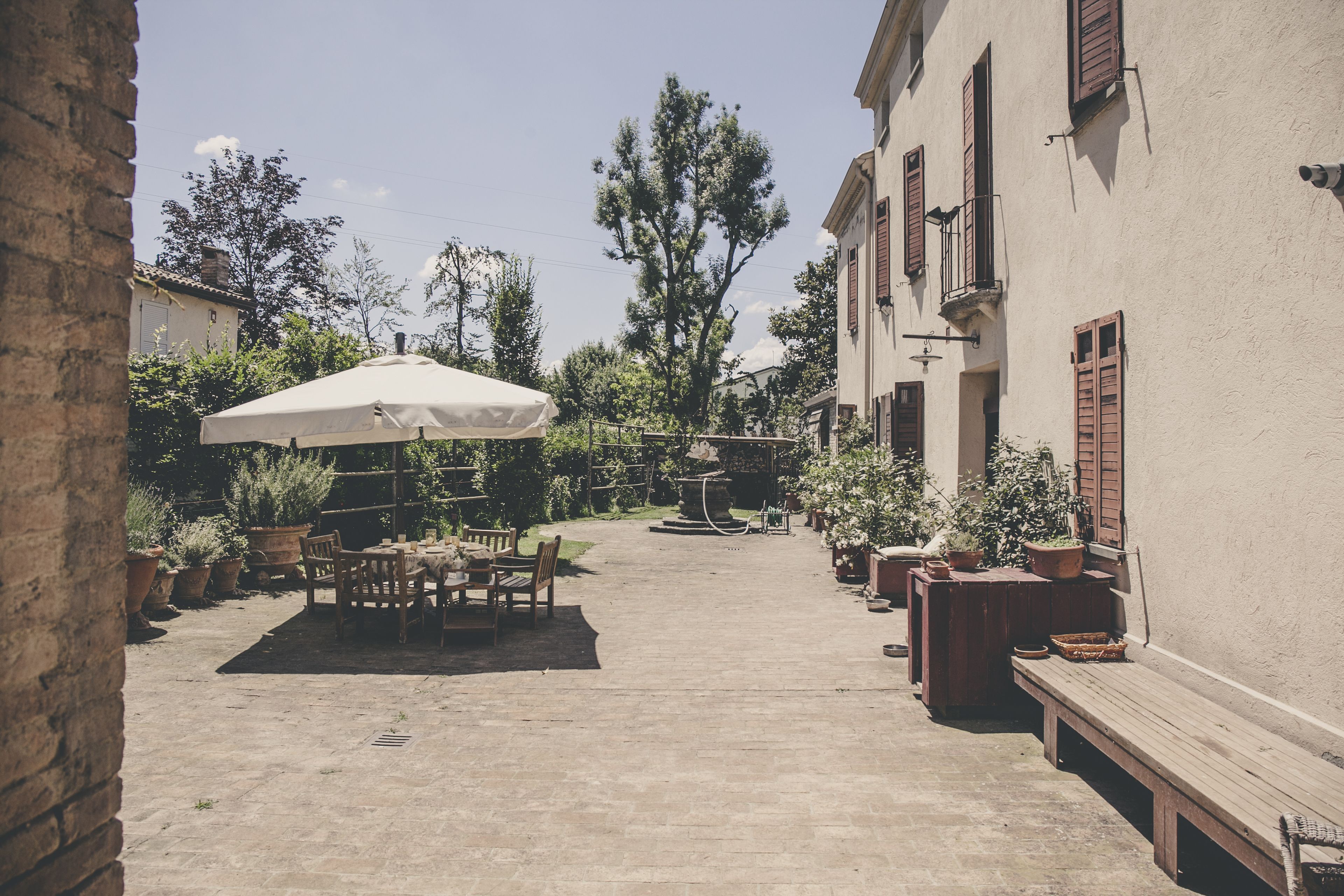 Une cour avec une table et des chaises sous un parasol blanc, entourée de plantes en pot et d’une fontaine.