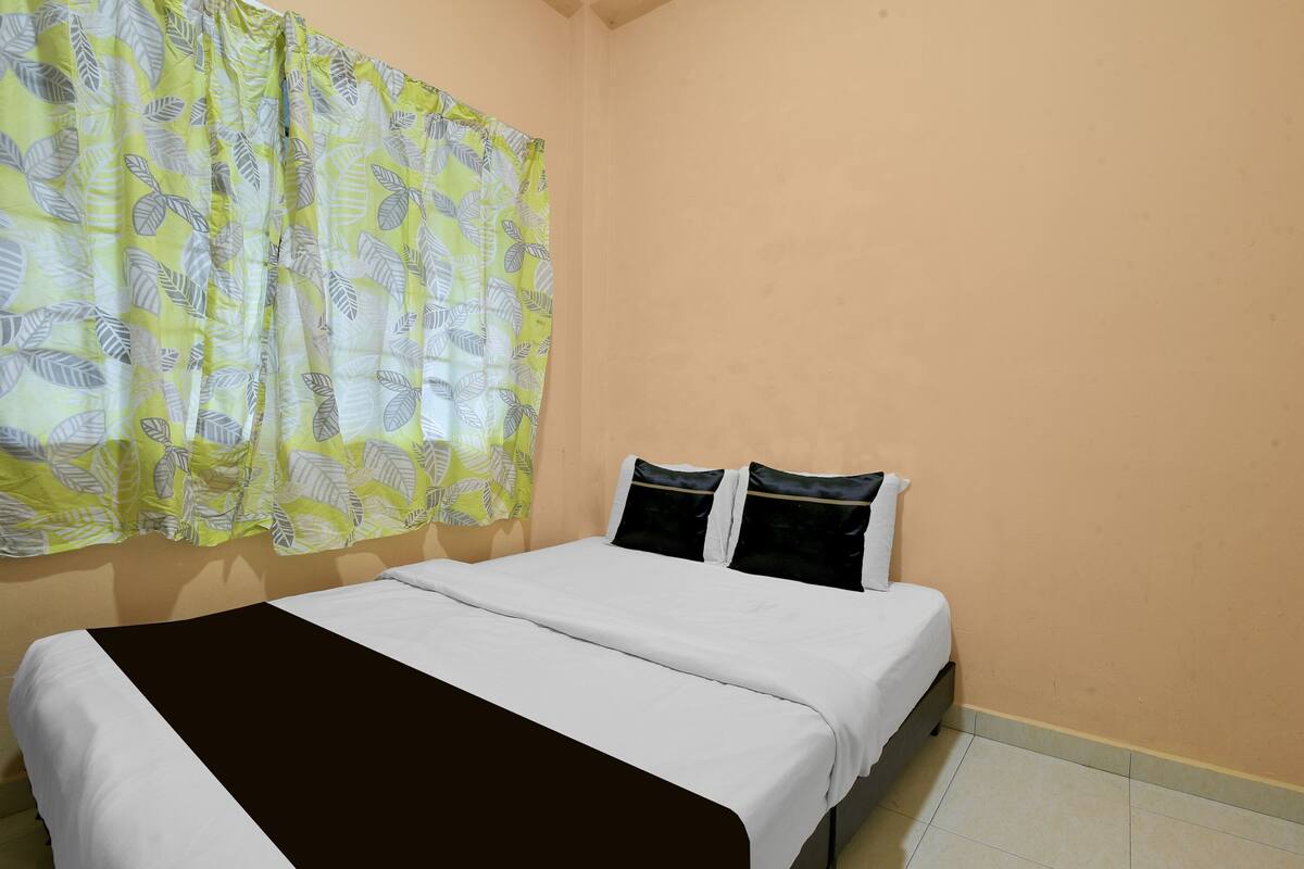 A room with a bed, a window with a patterned curtain, and a plain wall.