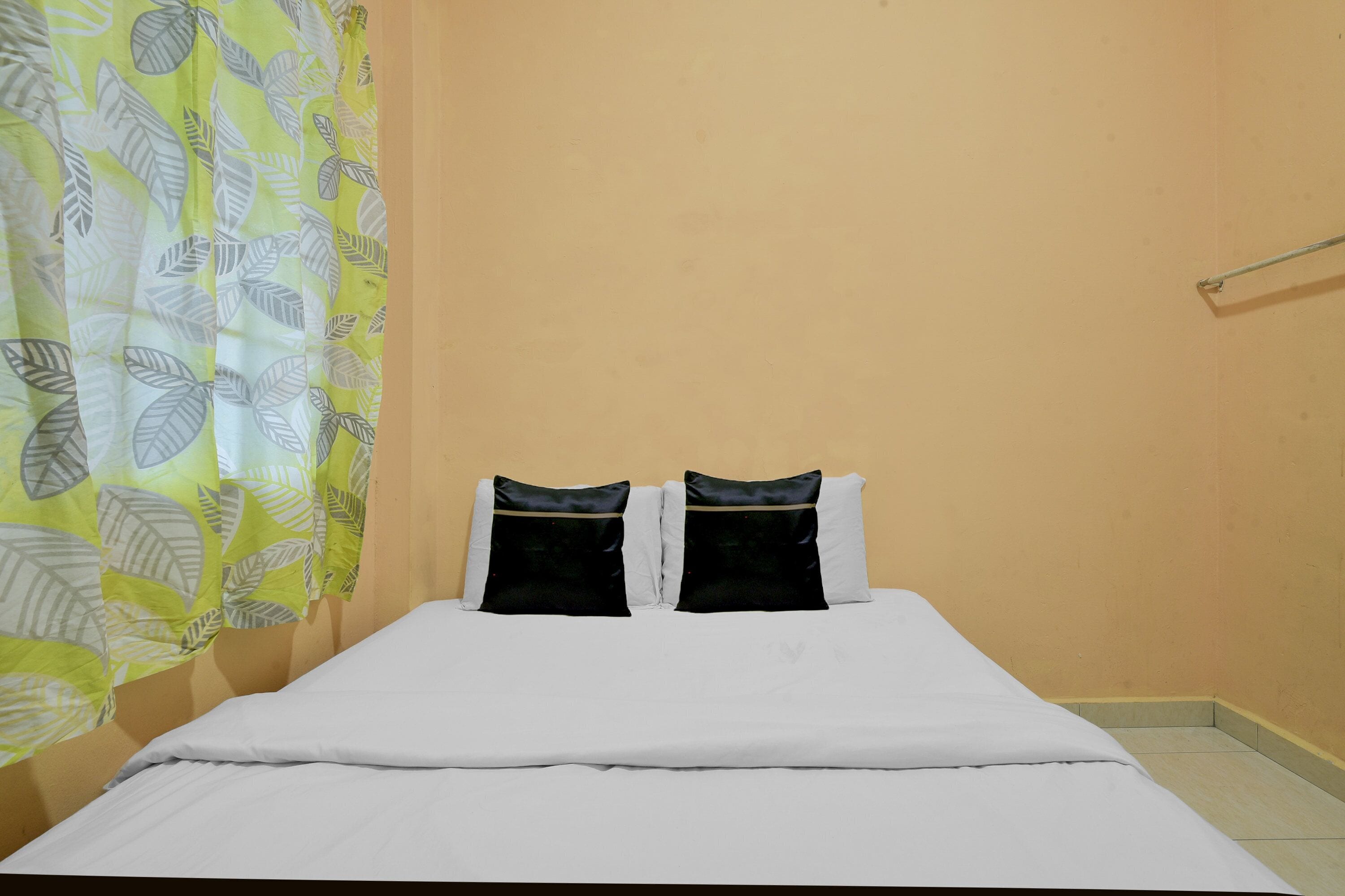 Un lit avec une literie blanche et deux oreillers noirs, un rideau jaune orné de motifs de feuilles et un mur de couleur beige.