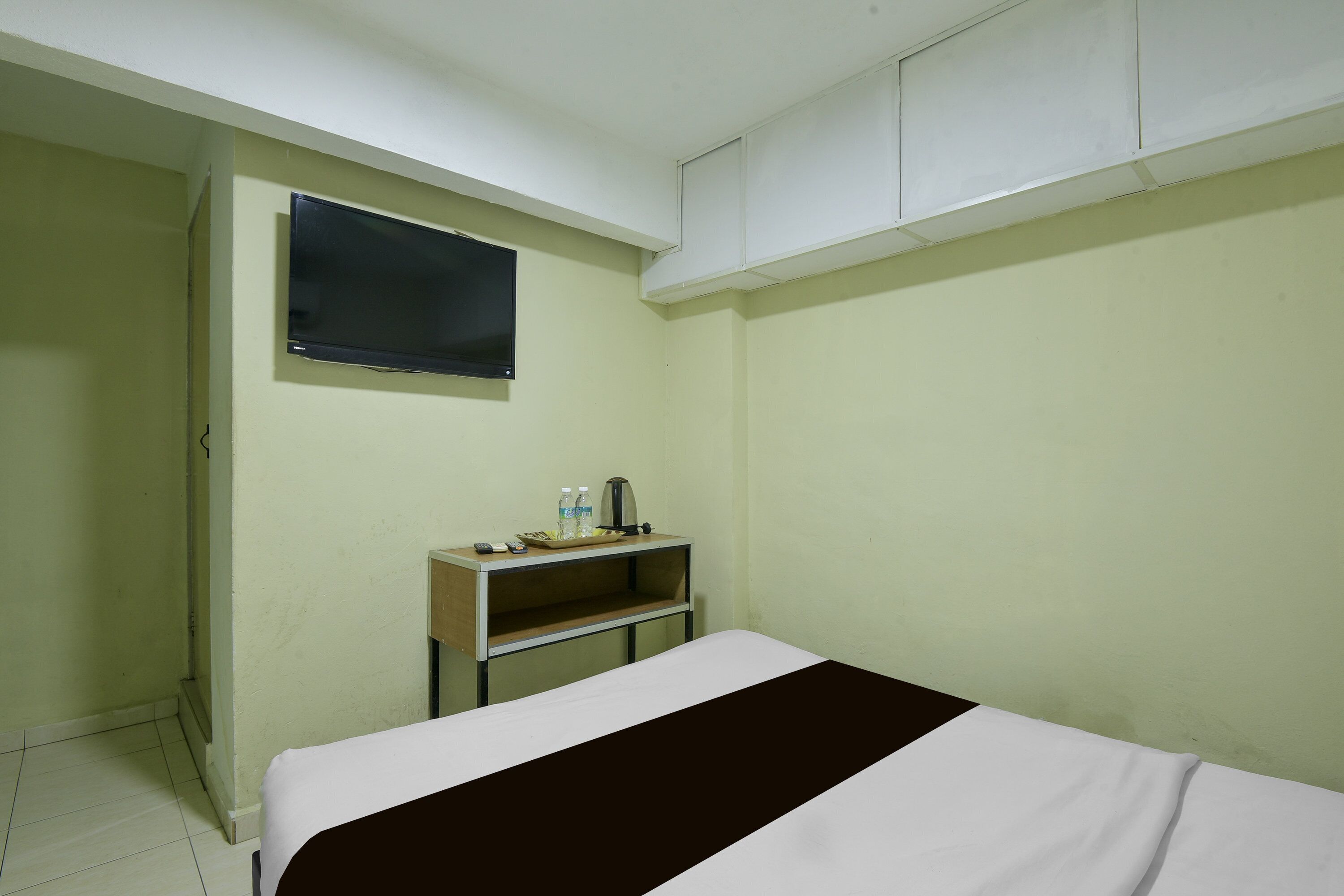 A room with a bed, a television mounted on the wall, a shelf with a kettle, and a water bottle.