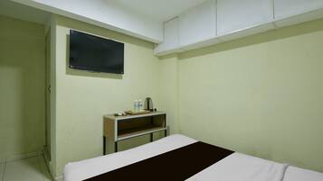 A room with a bed, a television mounted on the wall, a shelf with a kettle, and a water bottle.