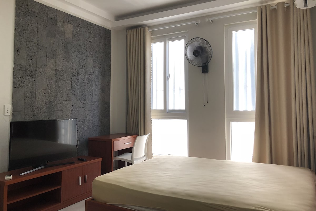 A hotel room with a bed, a TV on a wooden stand, a desk, a chair, a wall-mounted fan, and large windows with curtains.