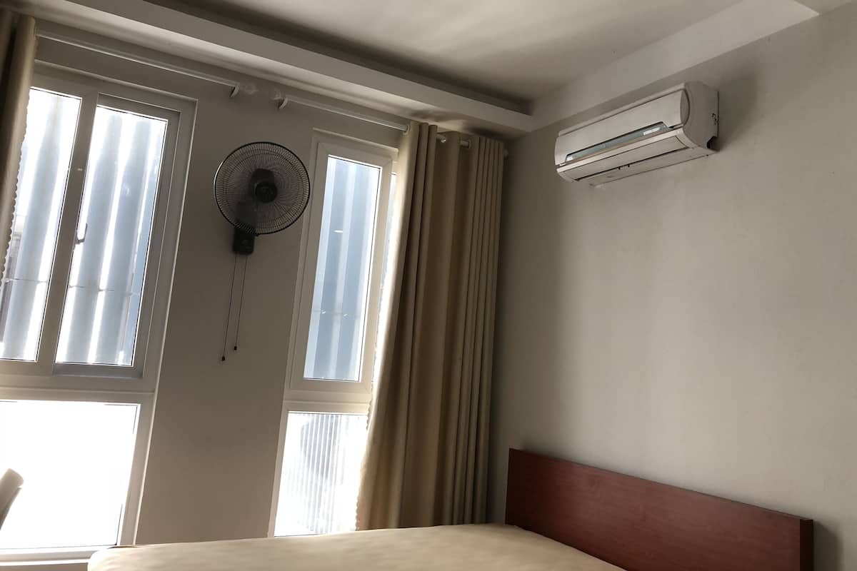 A hotel room with a bed, an air conditioner, a standing fan, and windows with curtains.