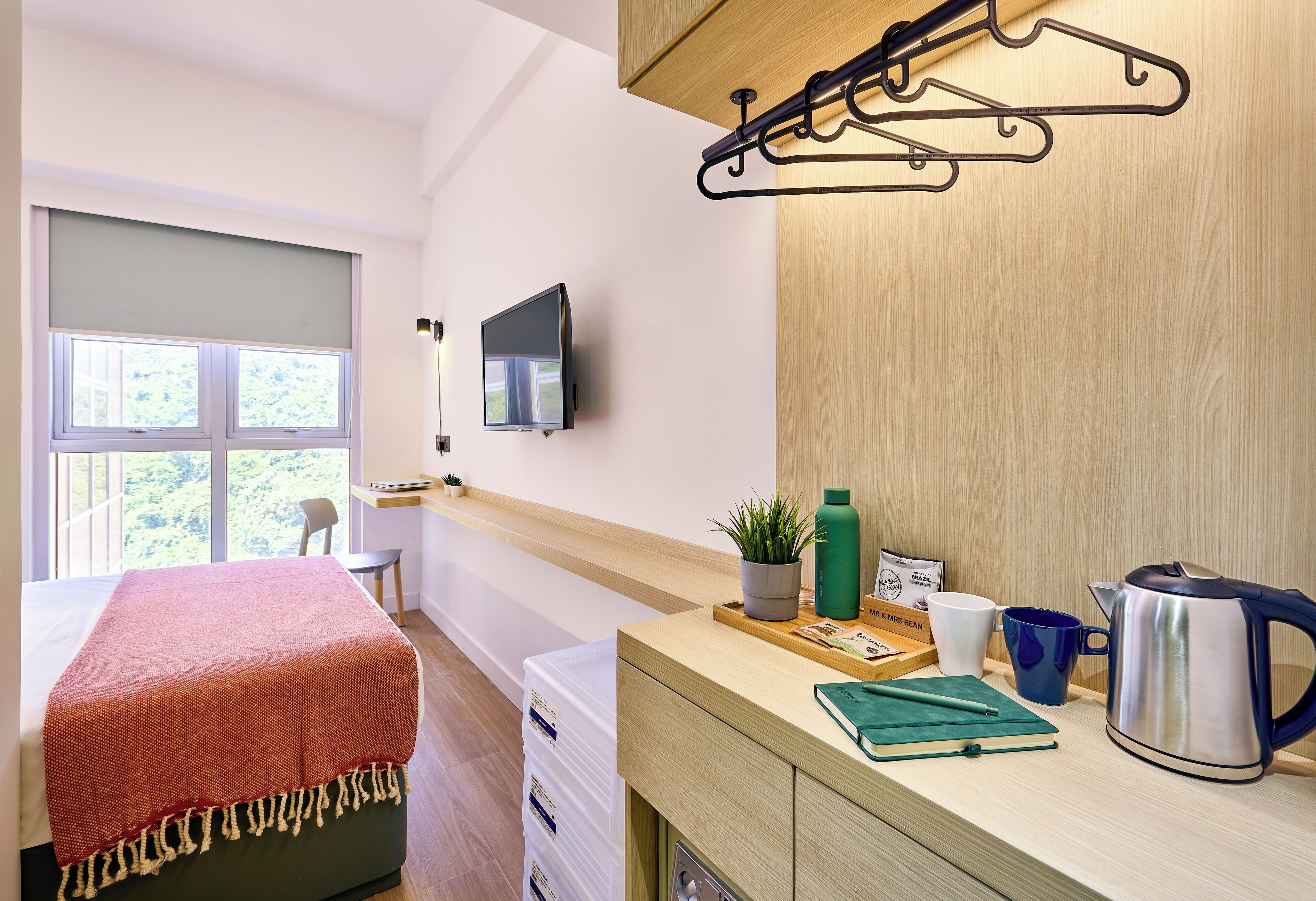 A hotel room with a bed, a TV mounted on the wall, a wooden desk with a kettle, a plant, and a coffee mug.