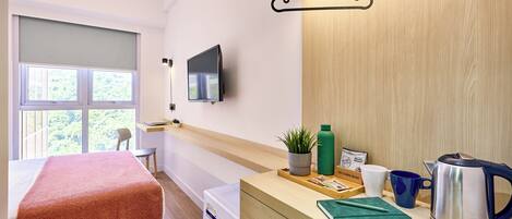 A hotel room with a bed, a TV mounted on the wall, a wooden desk with a kettle, a plant, and a coffee mug.