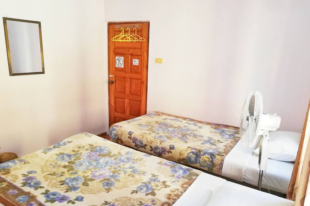 A room with two beds, each with floral bedding, a wooden door with a hanger, and a standing fan.