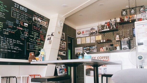 A modern cafe with a blackboard menu, a counter with a coffee machine, and a seating area with stools and a couch. - JJBro Hostel Guest House (Mokpo)