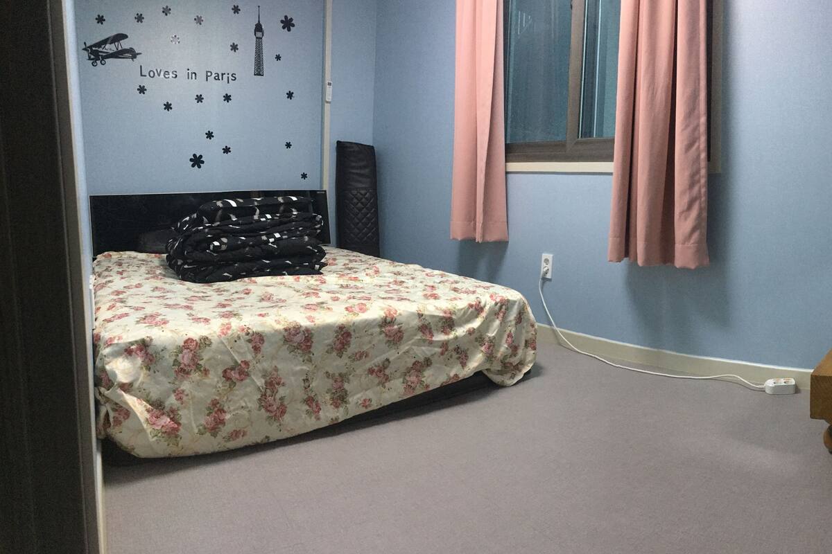 Une chambre avec un lit, une fenêtre ornée de rideaux roses, un motif décoratif mural représentant un paysage urbain, une table de chevet en bois et une poubelle.