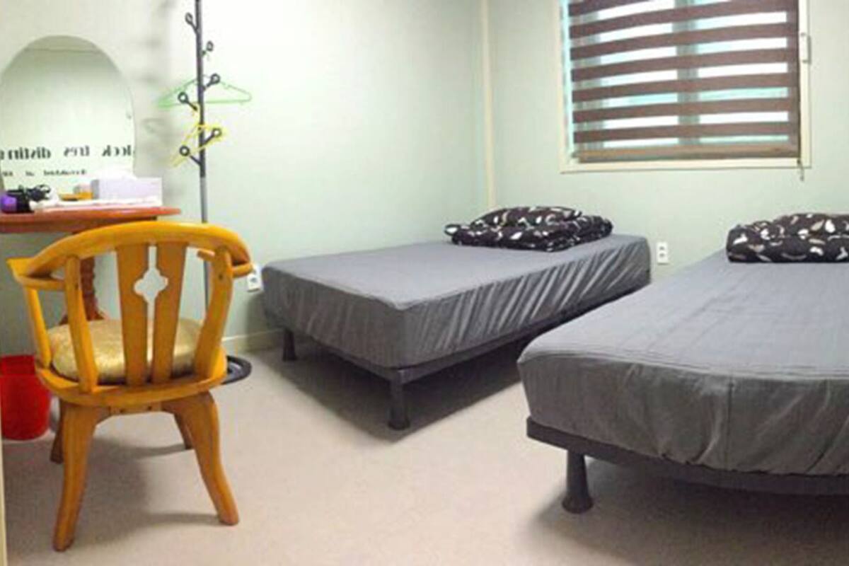 Une petite pièce comprenant des toilettes, une chaise en bois, deux lits simples et une fenêtre avec des stores.