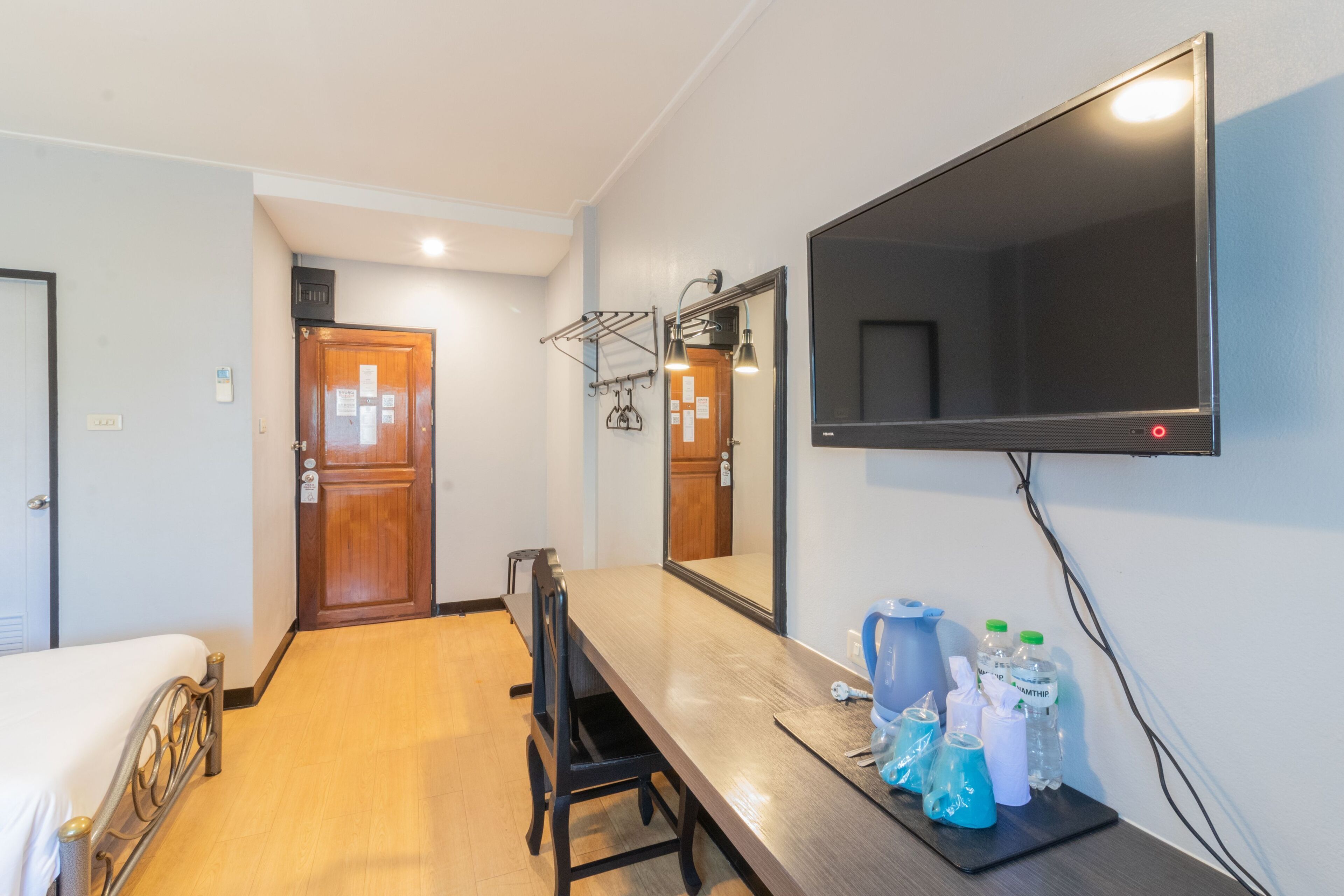 A hotel room with a bed, a wooden desk, a chair, a TV mounted on the wall, and a door with a mirror and towel rack.