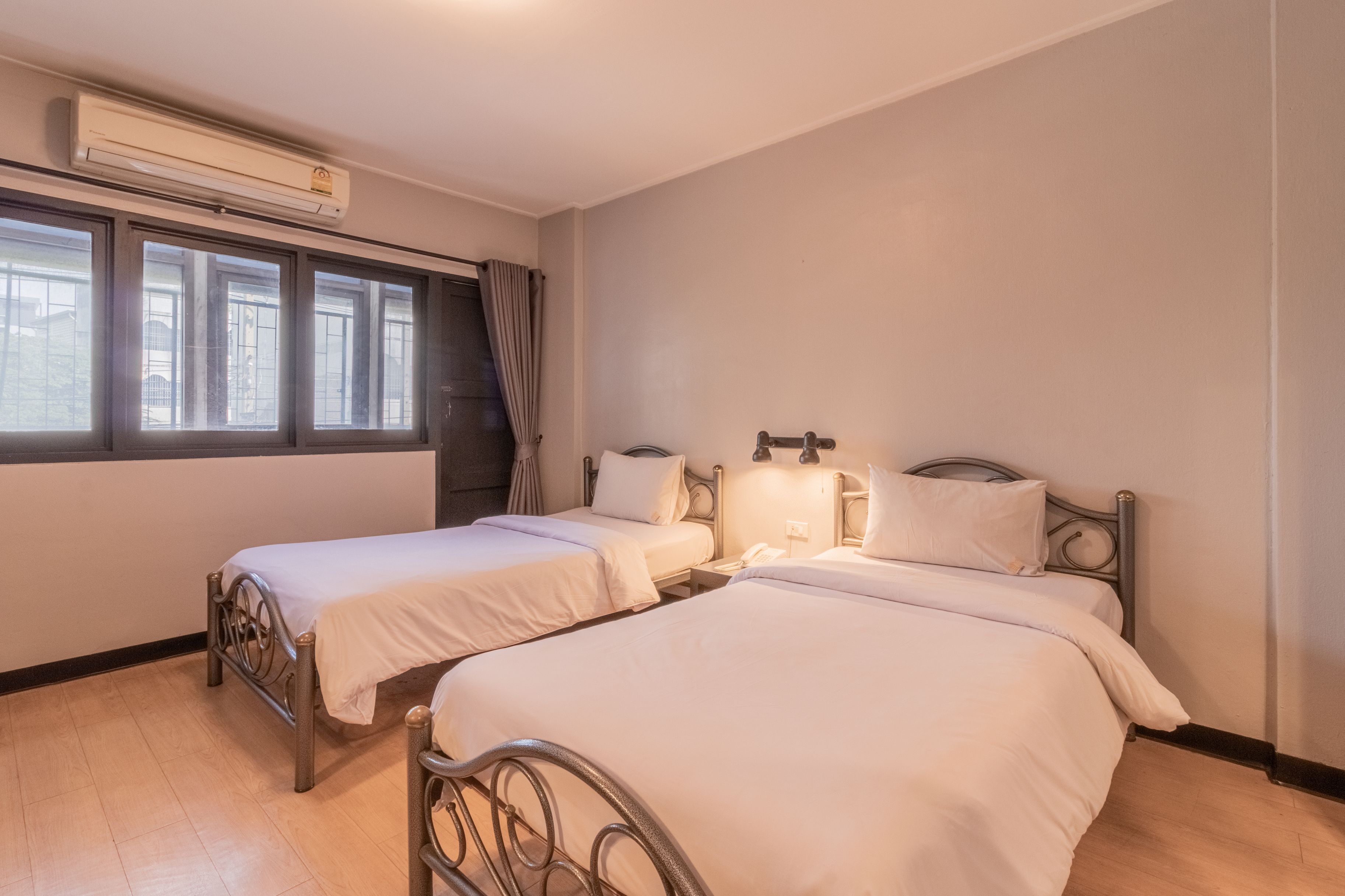 A room with two beds, an air conditioning unit, a window with curtains, and a nightstand with a lamp.