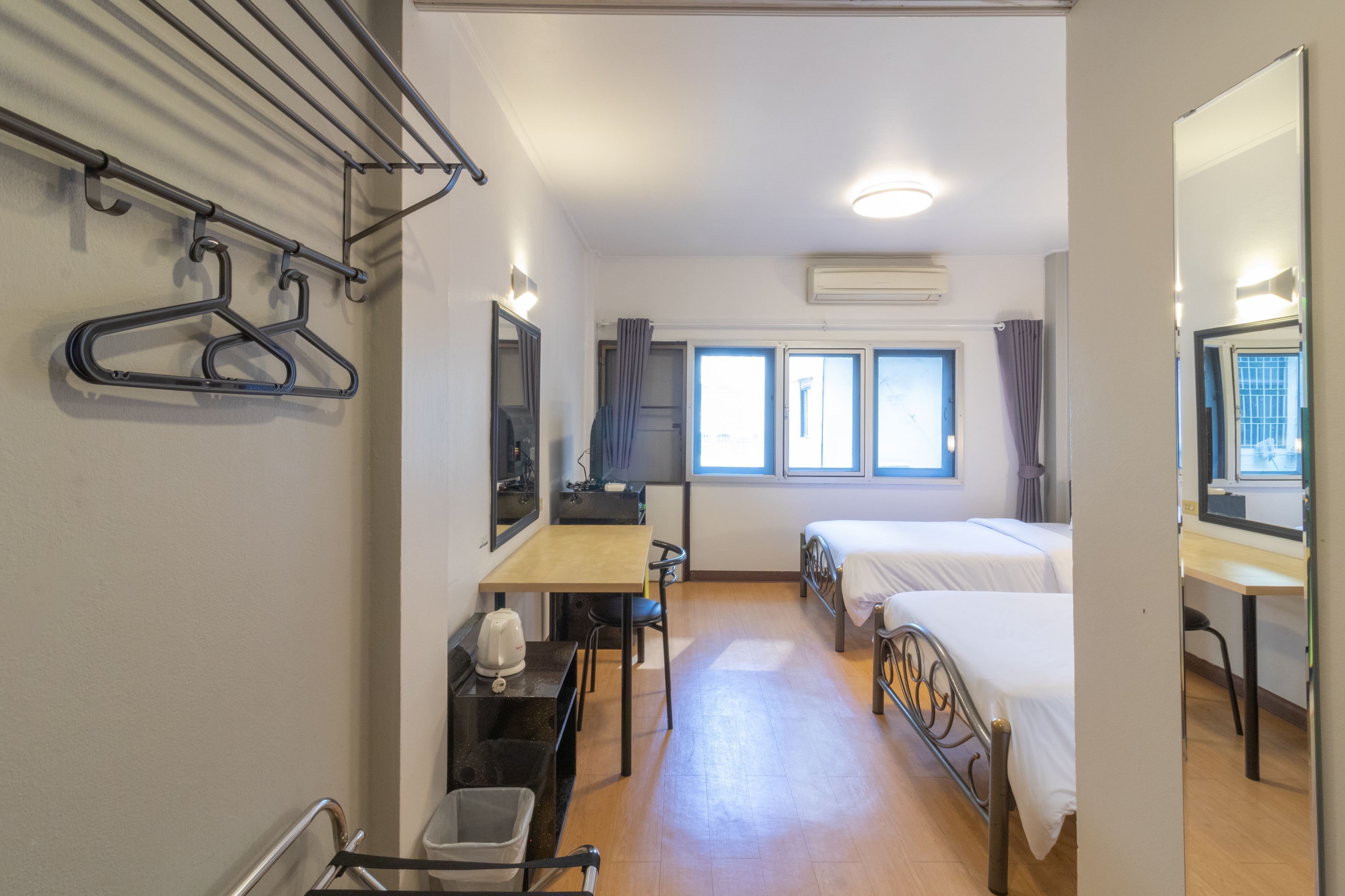 A hotel room with two beds, a desk, a chair, a small table, a kettle, and a drying rack with hangers.