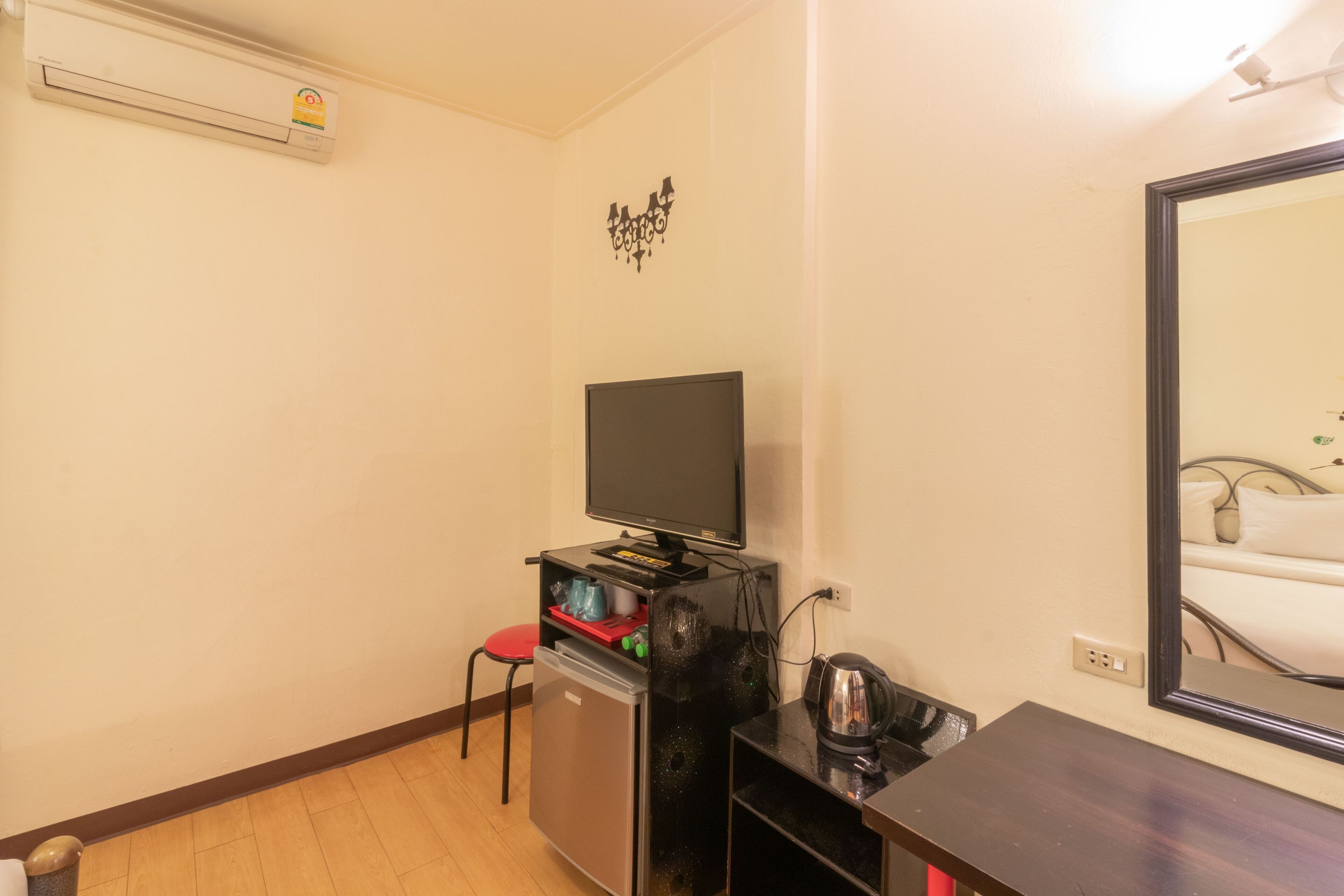A compact room with a flat-screen TV, a small refrigerator, a red chair, a black table, and a mirror reflecting a bed.