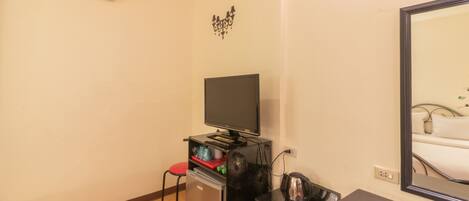 A compact room with a flat-screen TV, a small refrigerator, a red chair, a black table, and a mirror reflecting a bed.