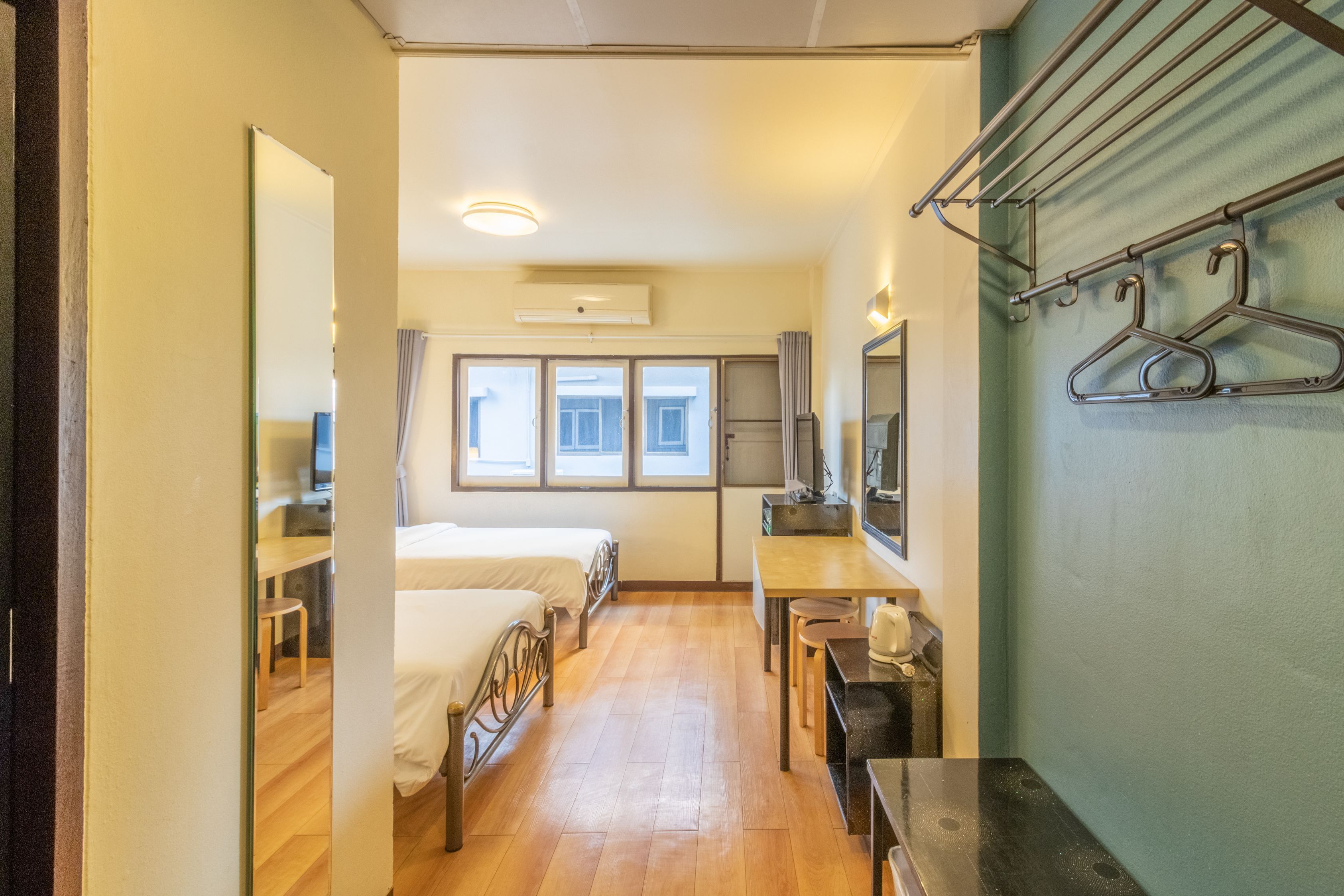A hotel room with a bed, a desk, a chair, and a mirror. There are windows with curtains, a hanging clothes rack, and an air conditioning unit on the wall.