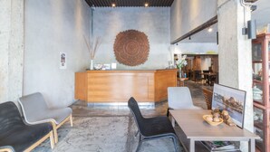 Uma área de recepção com balcão de madeira, duas cadeiras cinzas, uma cadeira preta, uma mesa com um recipiente de objetos decorativos e uma grande decoração circular na parede.