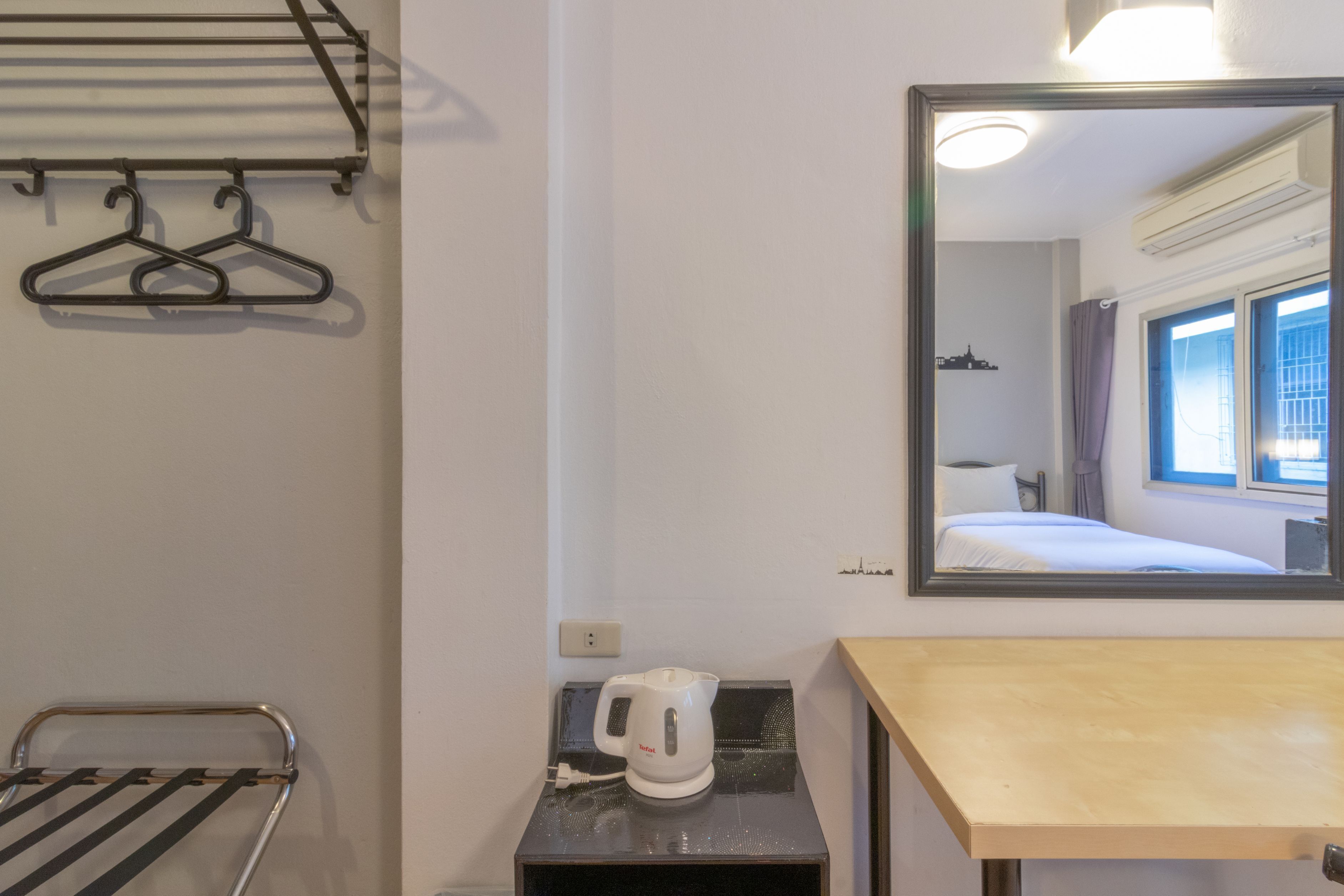 A hotel room with a bed, a mirror, a kettle, a towel rack, and a small table.
