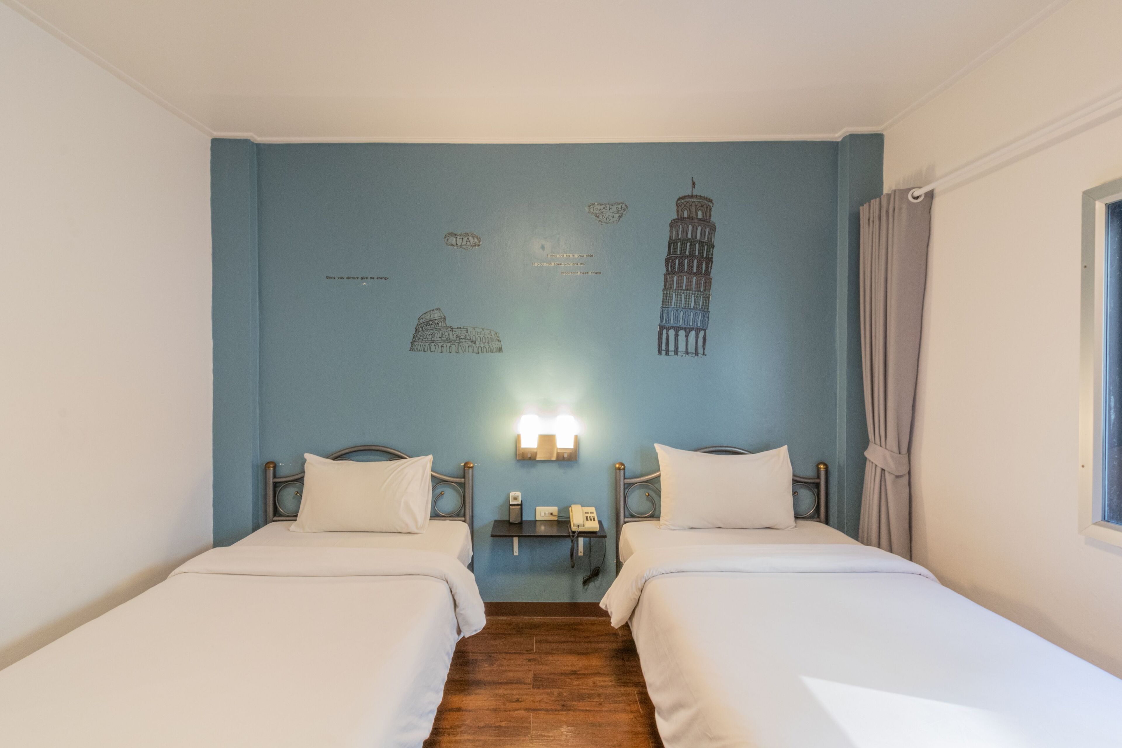 Ein Hotelzimmer mit zwei Betten, einem Nachttisch mit Telefon, einer Wand mit Zeichnungen des Kolosseums und des Schiefen Turms von Pisa sowie einem Fenster mit Vorhängen.