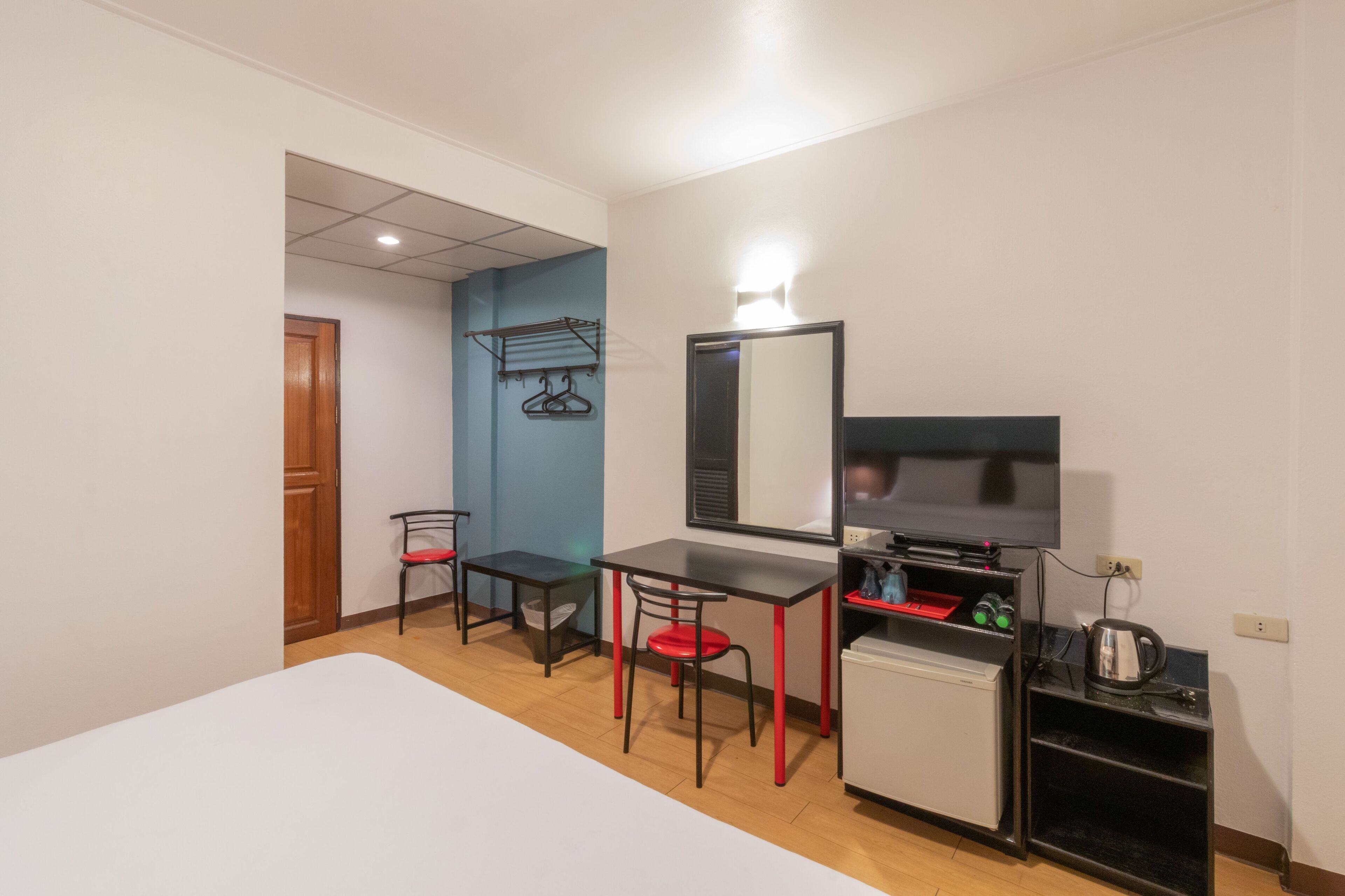 A compact hotel room with a bed, a small desk with a chair, a television, a mini-fridge, and a coat rack.