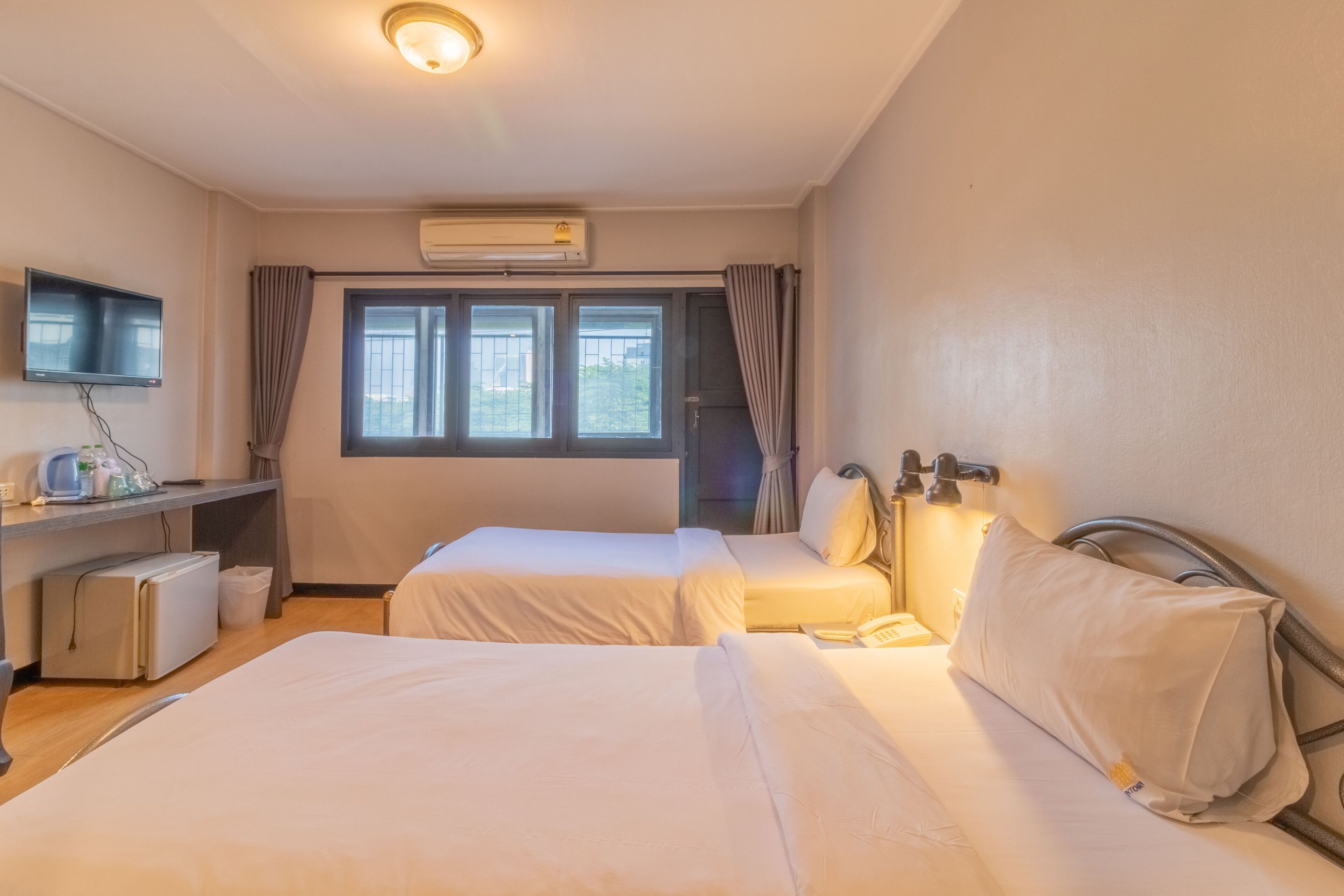 A hotel room with two beds, a desk with a computer, a TV, and an air conditioning unit.